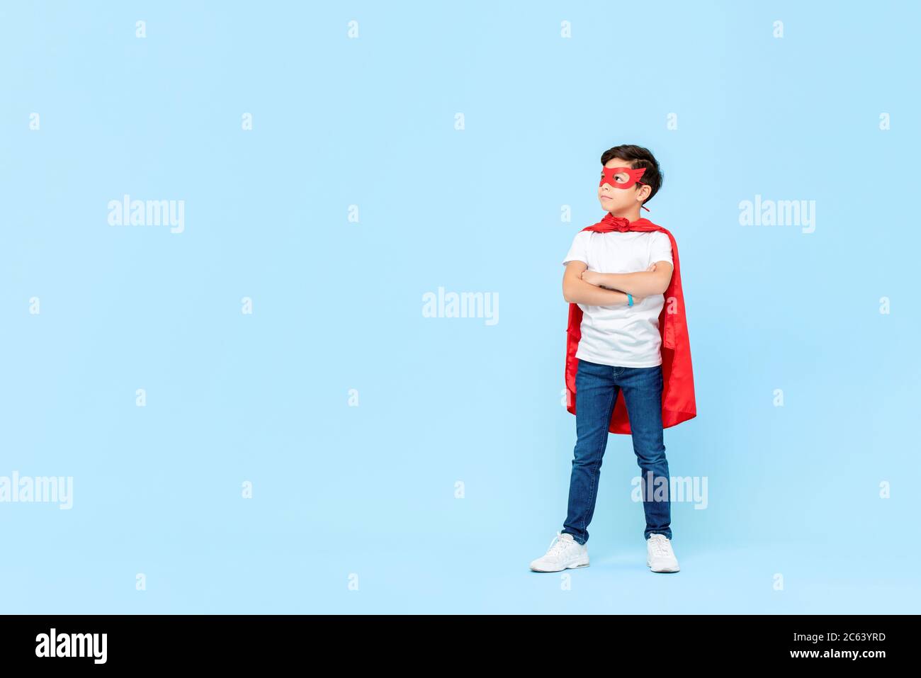Superhelden Junge in roter Maske und Umhang mit Armen gekreuzt Denken und Blick auf Kopie Raum beiseite isoliert hellblauen Hintergrund Stockfoto