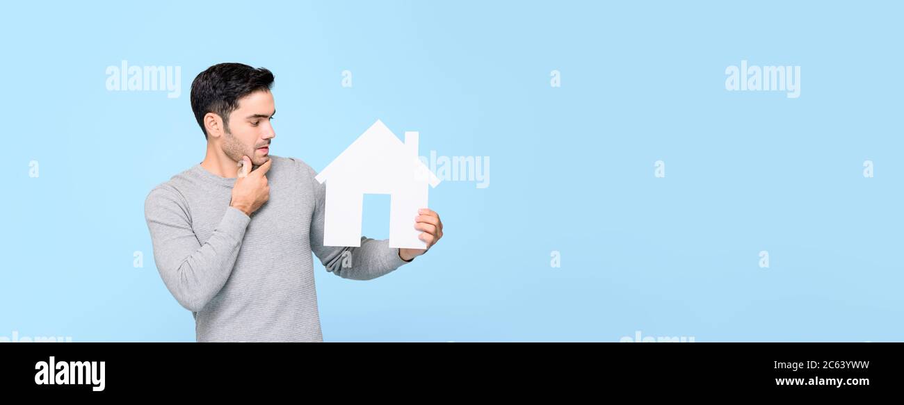 Junger Mann, der sich das Hausmodell ansieht und isoliert auf hellblauem Banner-Hintergrund mit Kopierraum denkt Stockfoto