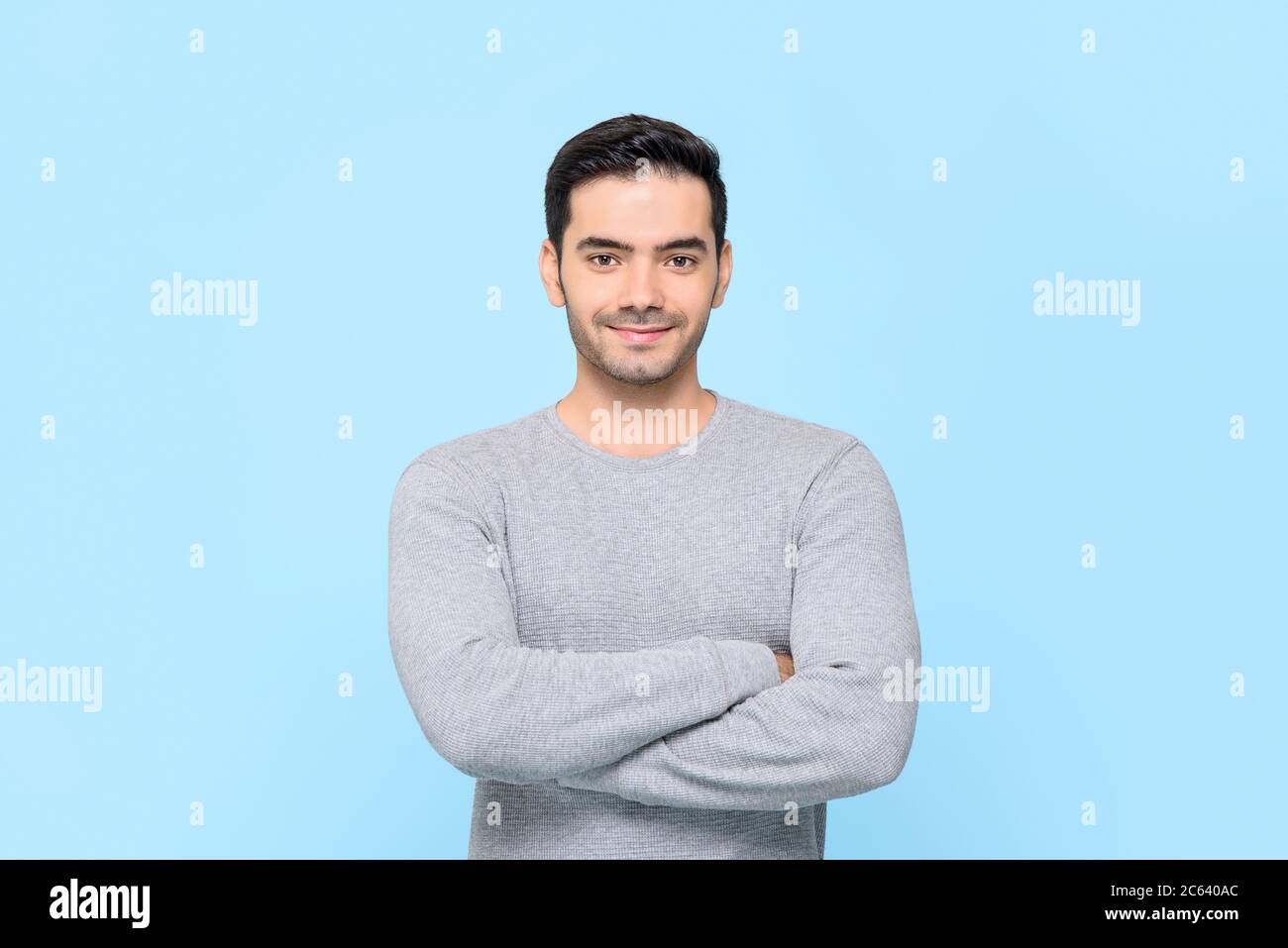 Porträt eines lächelnden freundlichen gutaussehenden Mannes in einfarbig grauem T-Shirt mit Armen, die isoliert in hellblauem Studiobakground gekreuzt sind Stockfoto