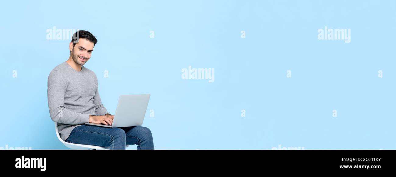 Lächelnder, hübscher Mann, der auf einem Stuhl sitzt, der auf einem Laptop-Computer arbeitet, der auf hellblauem Banner-Hintergrund mit Kopierraum isoliert ist Stockfoto