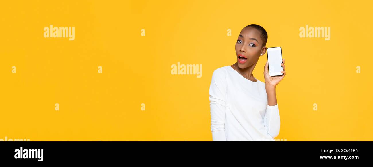Überrascht schöne afroamerikanische Frau hält Handy mit leerem Bildschirm auf isolierten gelben Banner Hintergrund mit Kopieplatz Stockfoto