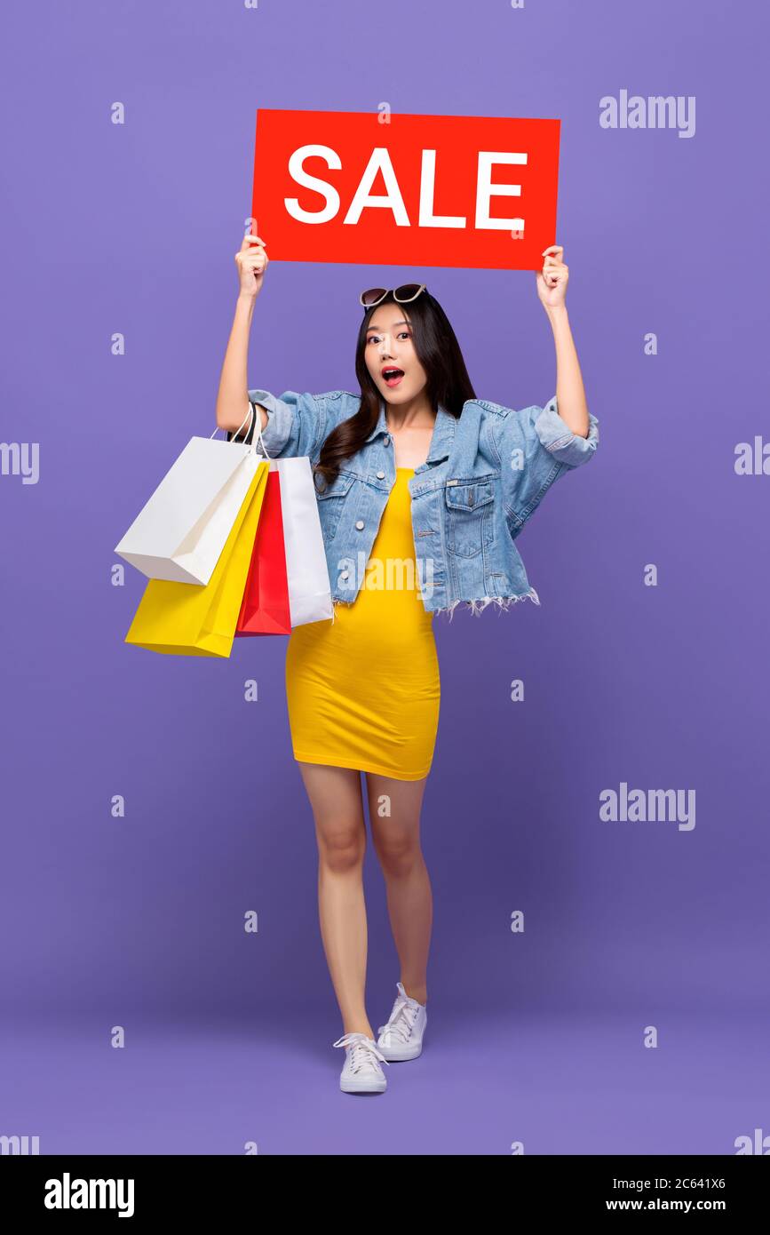 Überrascht modische asiatische Frau trägt colofrul Einkaufstaschen Erhöhung roten Verkauf Zeichen isoliert auf lila Hintergrund Stockfoto