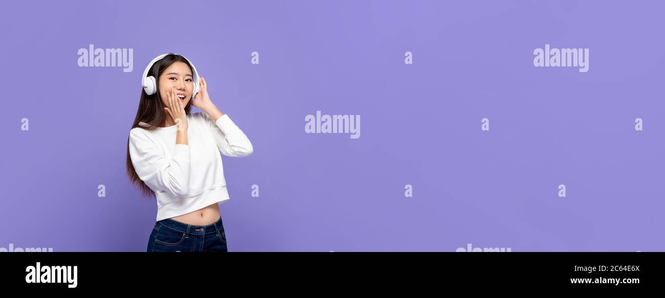 Junge hübsche asiatische Frau lächelt und deckt Mund, während Musik neben Kopie Raum auf isolierten lila Banner Hintergrund hören Stockfoto