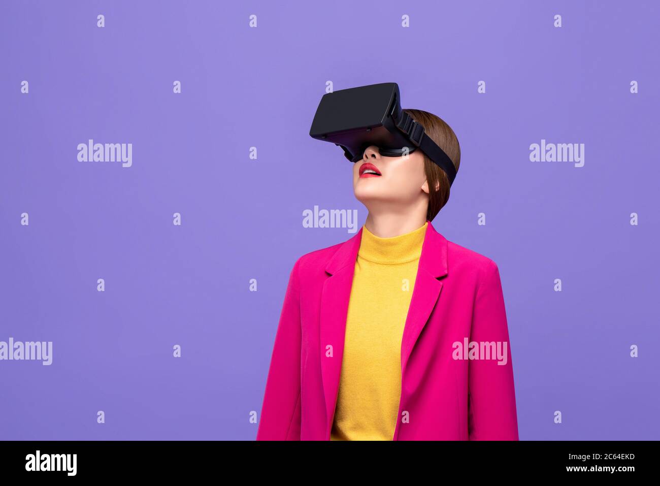 Frau in farbenfroher Kleidung, die den Kopf hebt und 3D-Simulationsvideos aus der virtuellen Realität oder VR-Brillen auf purpurem Hintergrund ansieht Stockfoto