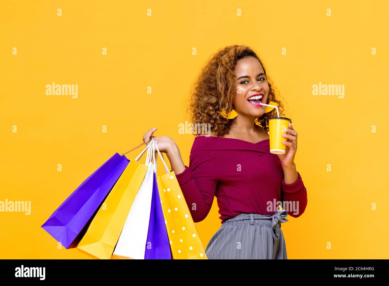 Portrait von fröhlichen jungen attraktiven afroamerikanischen Frau hält Einkaufstaschen während trinken in isolierten Studio gelben Hintergrund Stockfoto