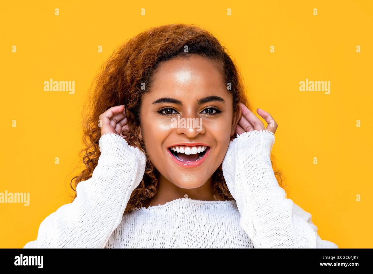 Nahaufnahme Porträt einer fröhlichen jungen afroamerikanischen Frau lächeln, während sie ihre Ohren mit beiden Händen in isolierten Studio gelben Hintergrund berühren Stockfoto