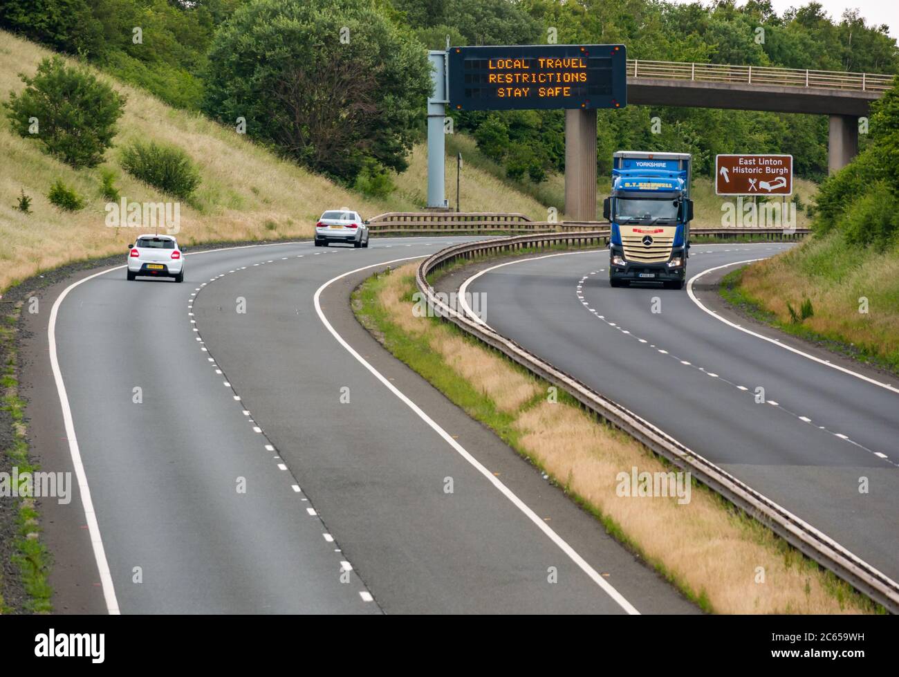 East Lothian, Schottland, Großbritannien, 7. Juli 2020. Covid-19 neue Meldung auf A1-Gantry: Die dritte Version der Pandemie-Meldung erscheint auf der über-Gantry in der Nähe von Haddington, die lautet "Lokale Reisebeschränkungen sicher bleiben", da Schottland in Phase 3, die nächste Woche erwartet wird, eingeht. Der Verkehr auf der zweispurigen Fahrbahn ist viel schwerer als in den letzten Monaten zu sehen war Stockfoto