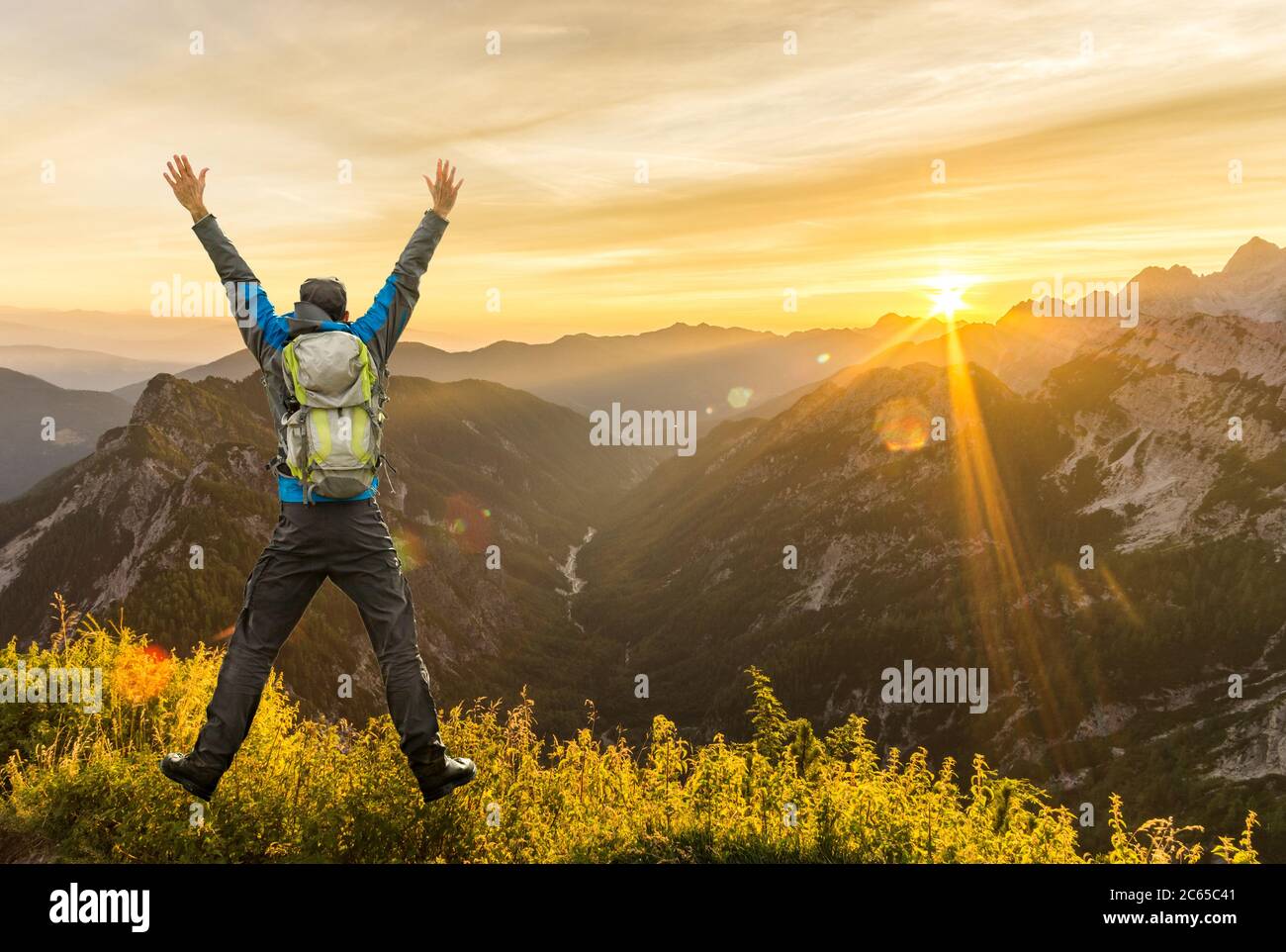 Wandermann mit Rucksack springen mit erhobenen Armen auf Berg. Erstaunliche Sonnenaufgang Hintergrundbeleuchtung mit schönen Linsen Fackeln und Sonnenstrahlen. Die Julischen Alpen, Triglav Stockfoto