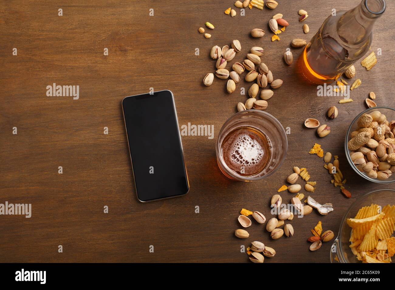 Smartphone auf Holztisch, neben einem Glas Bier mit Schaum, Flasche, Nüsse, Chips in Glasplatten Stockfoto