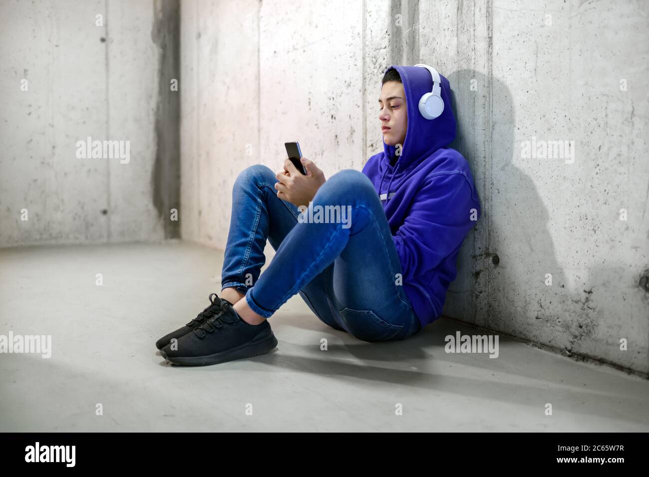 Kleiner Teenager-Junge mit Kopfhörern, der mit seinem Handy chattet, während er in einem blauen Hoodie und Jeans auf dem Betonboden in einer U-Bahn oder einem Korridor sitzt Stockfoto