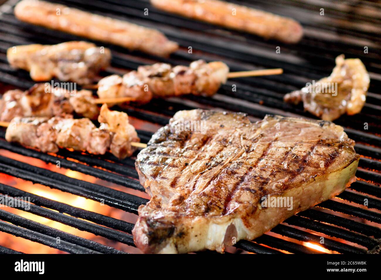 Grillen eines florentinischen Rindersteaks auf einem Grillfeuer mit Kebabs und Würstchen in Nahaufnahme mit Fokus auf das Steak über den heißen Kohlen Stockfoto