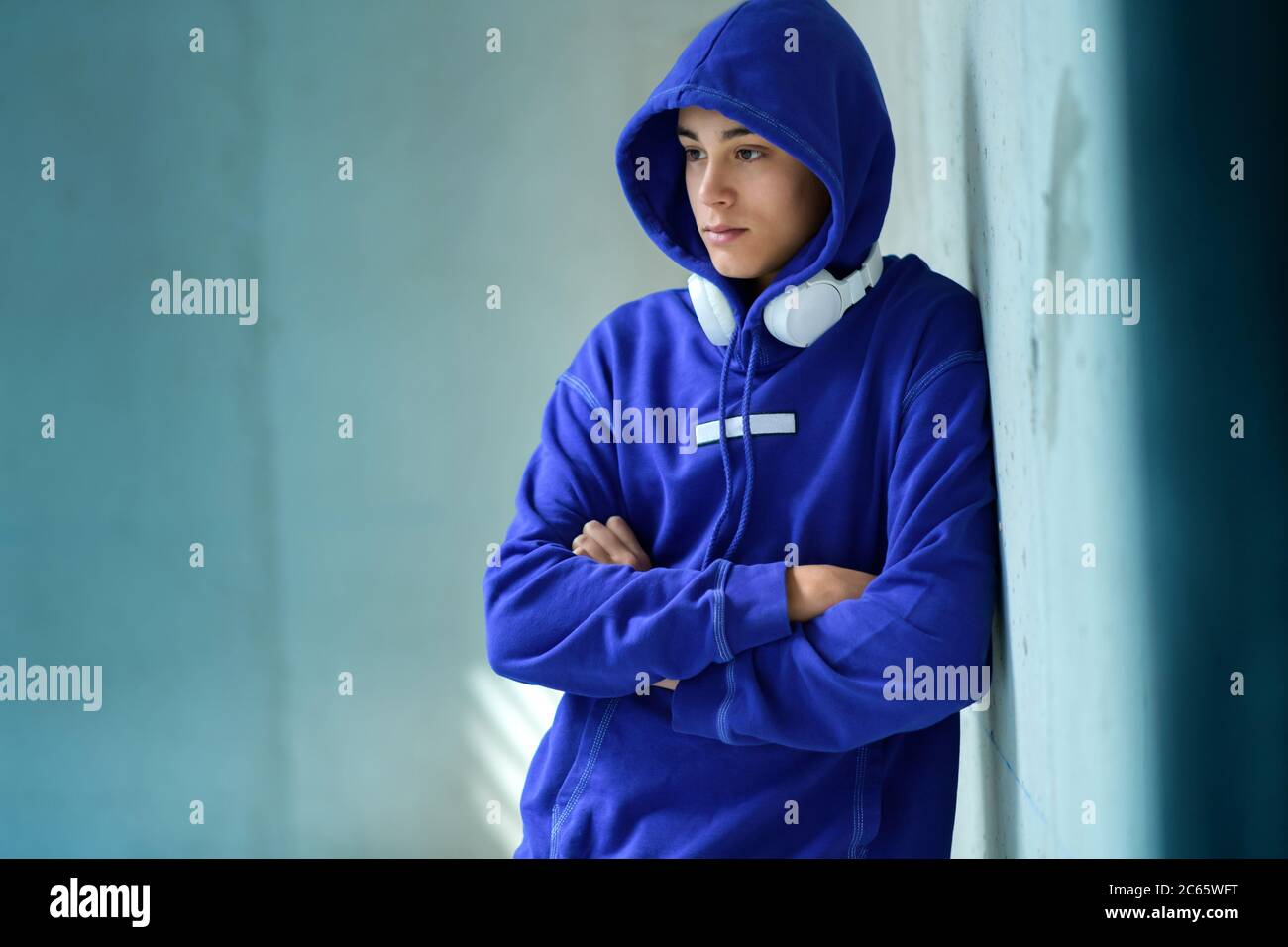 Nachdenklicher ernsthafter Teenager-Junge in einem blauen Hoodie, der mit gefalteten Armen auf einer blau getönten Wand in einem Sonnenstrahl steht und mit einem Stift zur Seite schaut Stockfoto