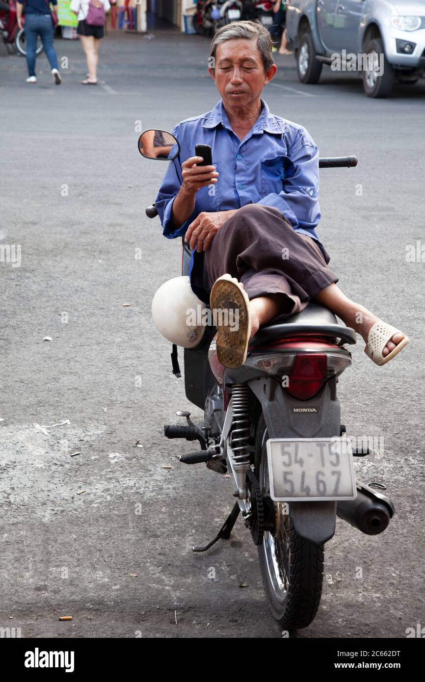 Vietnamesischer Mann in blauem Hemd entspannt auf seinem Motorrad auf der Seite der Straße mit Beinen gekreuzten Messaging auf seinem Handy. Stockfoto