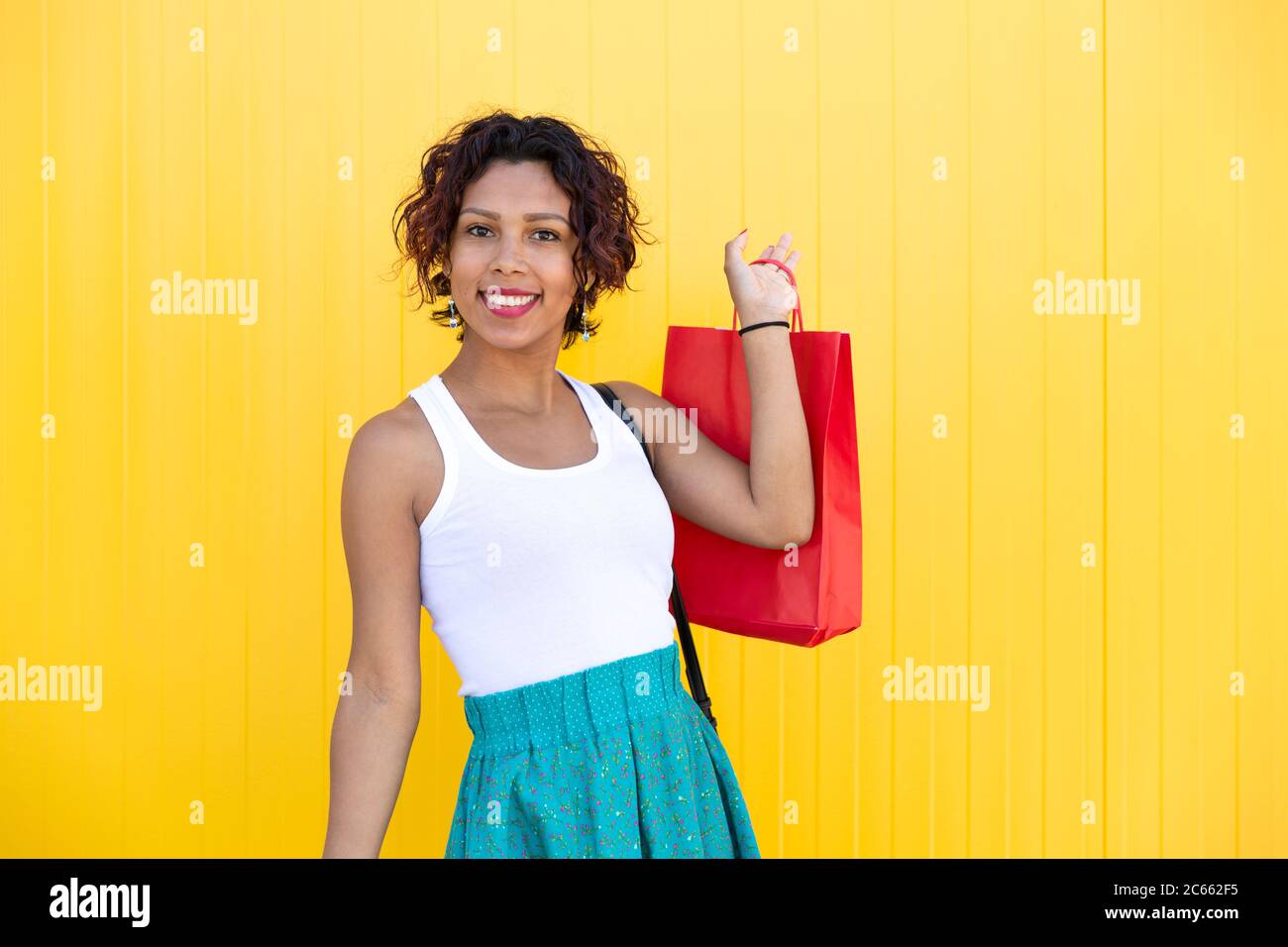 Junge Frau mit brauner Haut lächelt, als sie eine Einkaufstasche an einer gelben Wand zeigt. Leerzeichen für Text. Shopping, Geschenke und Verkaufskonzept. Stockfoto