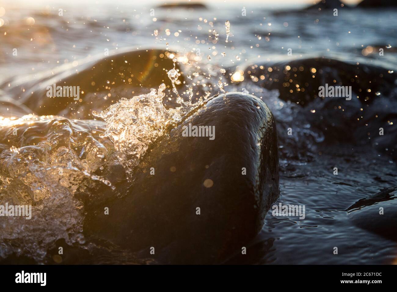 Nahaufnahme von Meereswellen, die auf einer nassen Küste brechen Steine Sommer Sonnenuntergang, Wasserspritzer, Sonnenstrahlen, Reflexionen auf Wassertropfen. Stockfoto