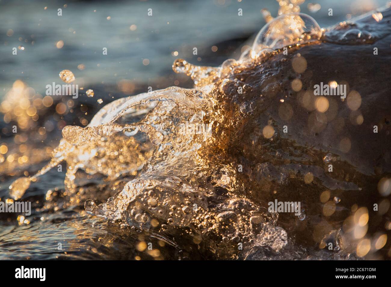Nahaufnahme von Meereswellen, die auf einer nassen Küste brechen Steine Sommer Sonnenuntergang, Wasserspritzer, Sonnenstrahlen, Reflexionen auf Wassertropfen. Stockfoto
