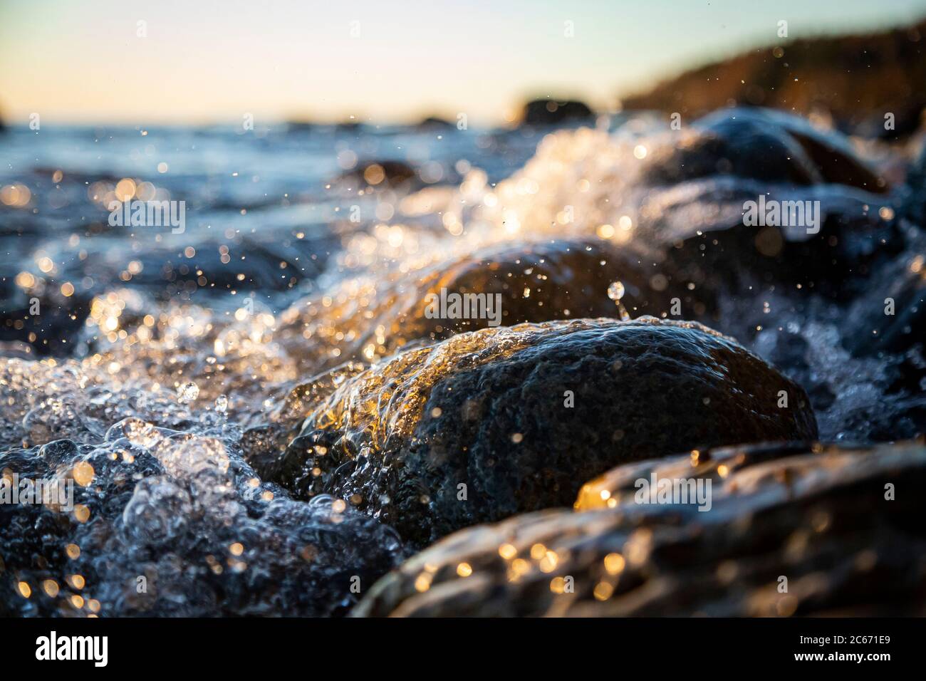 Nahaufnahme von Meereswellen, die auf einer nassen Küste brechen Steine Sommer Sonnenuntergang, Wasserspritzer, Sonnenstrahlen, Reflexionen auf Wassertropfen. Stockfoto