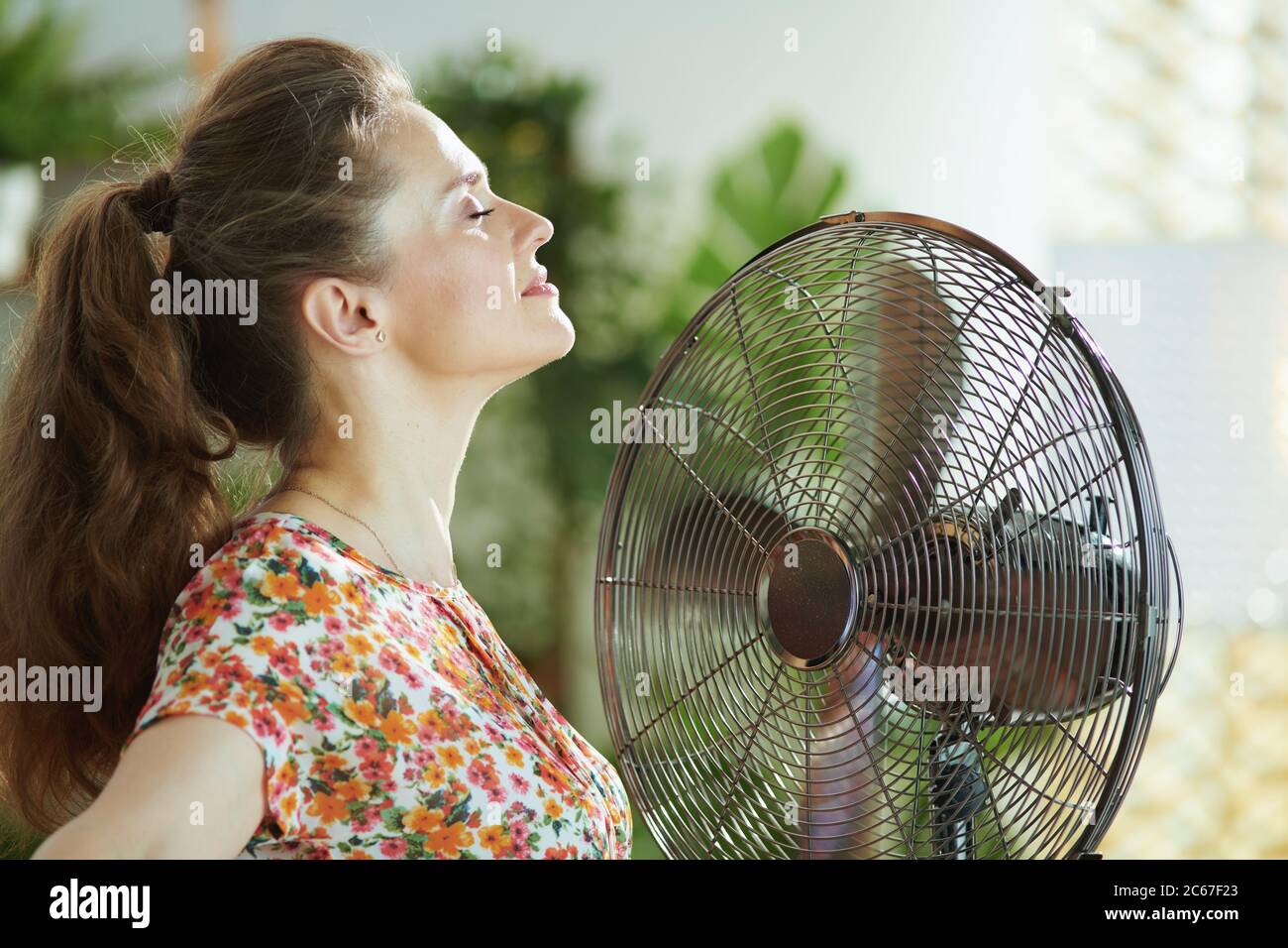 Sommerhitze. Entspannte junge Hausfrau in floraler Bluse im modernen Haus in sonnigen heißen Sommertag fächert sich. Stockfoto