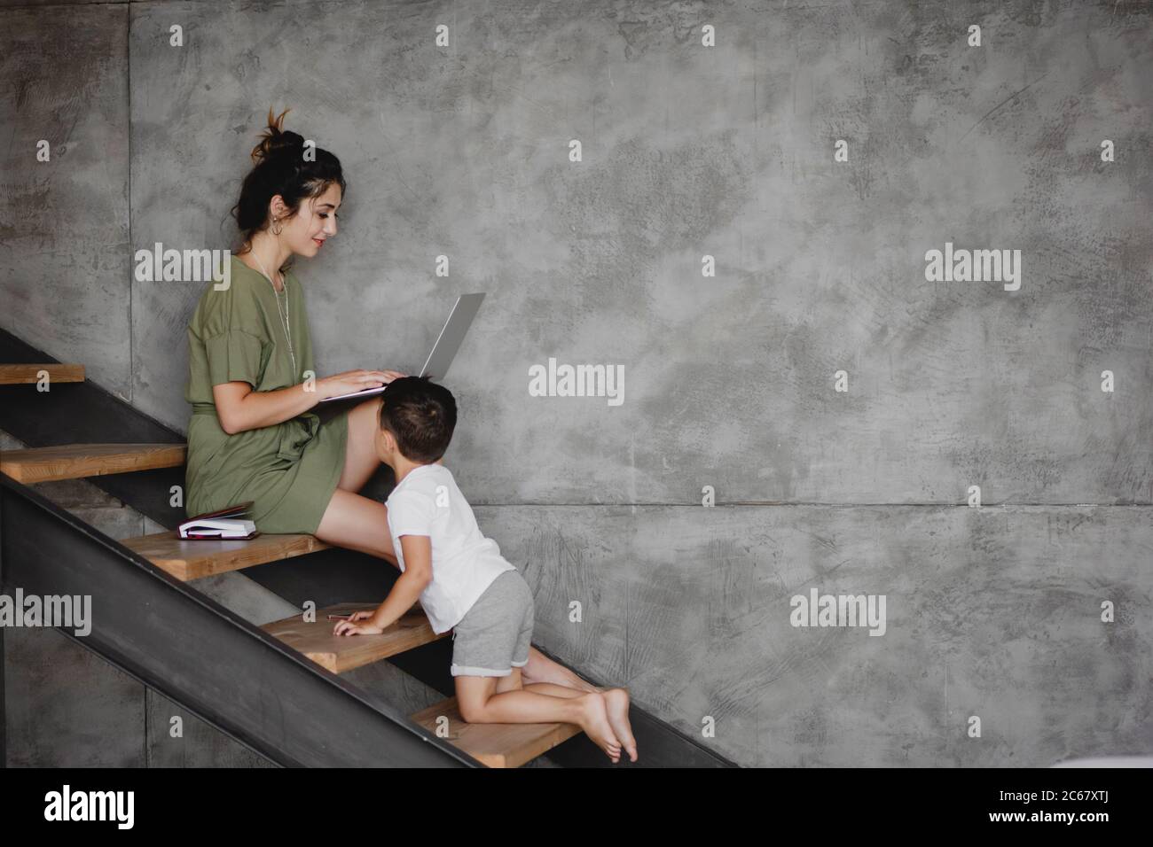 Junge Mutter sitzt auf der Treppe von zu Hause auf Laptop mit ihrem kleinen Sohn arbeiten. Stockfoto