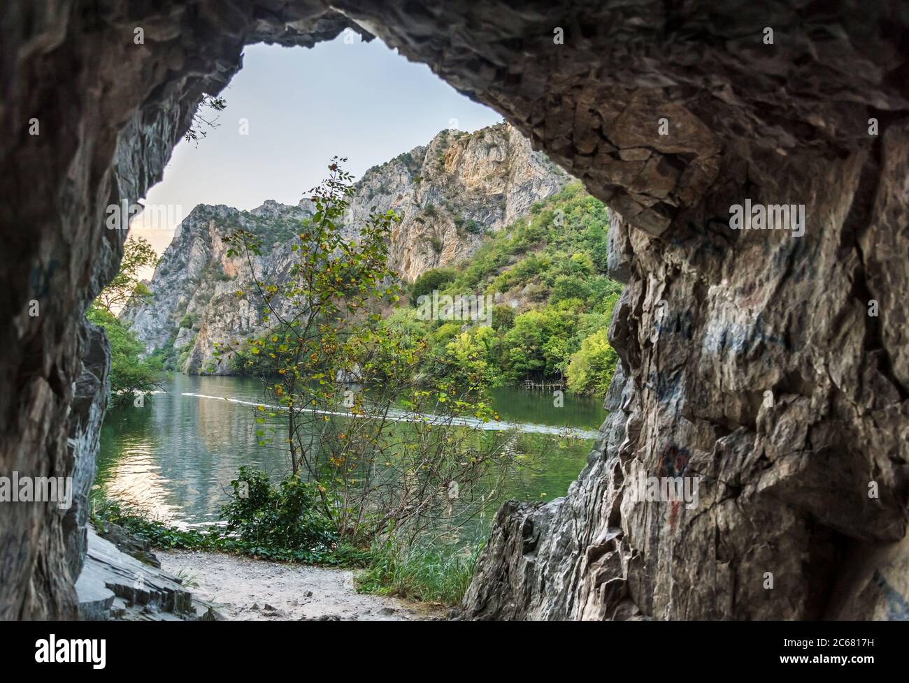 Ein Loch in der Bergseite als felsiger Weg windet sich entlang der Schlucht, und ein Boot driftet entlang des Flusses Treska und ruhigen Gewässern Matka See dazwischen Stockfoto
