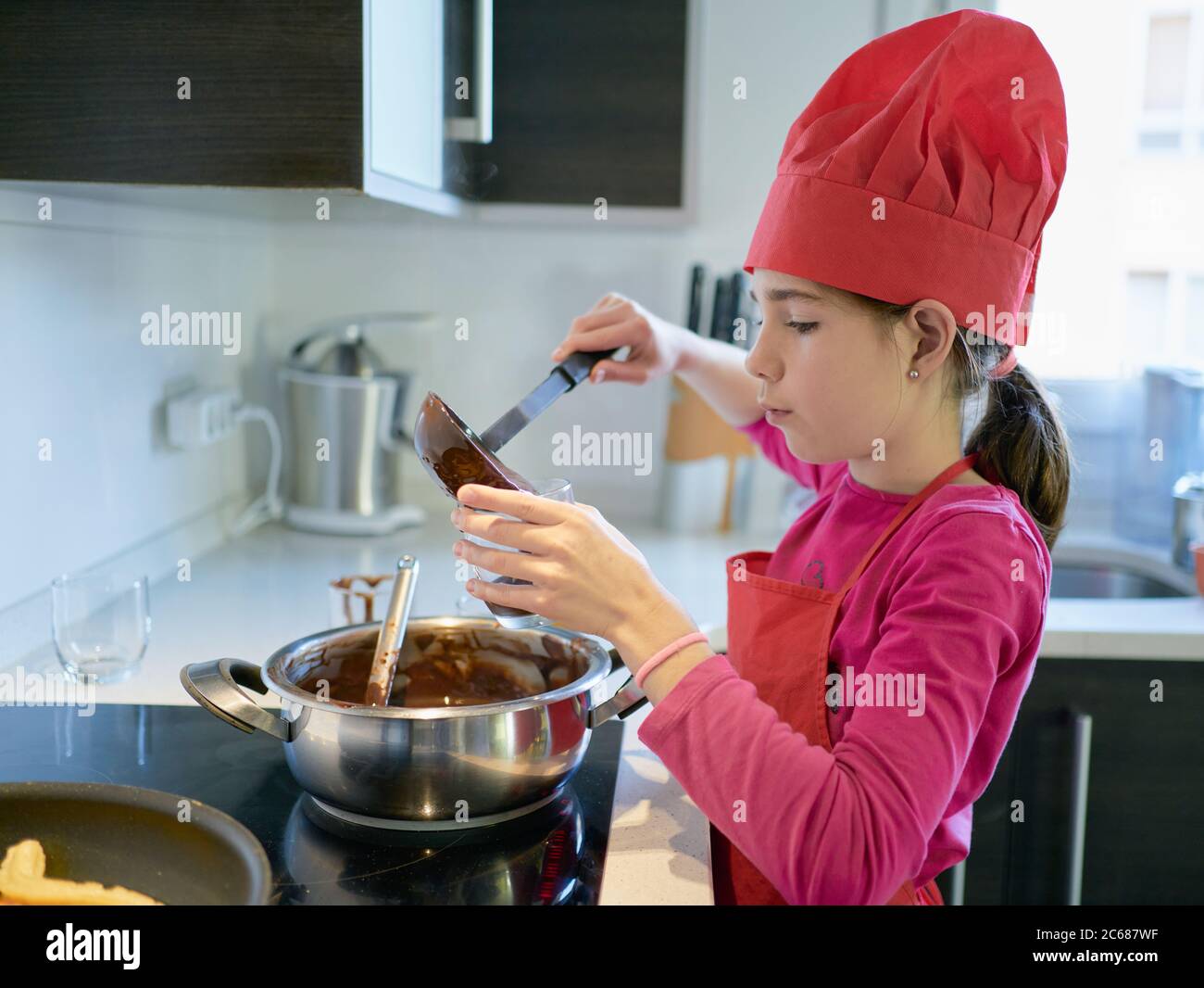 Mädchen in Kappe Gießen Schokoladensauce in Tasse während der Vorbereitung Dessert in der Küche Stockfoto Mädchen in Kappe Gießen Schokoladensauce in Tasse während der Vorbereitung Dessert in der Küche Stockfoto