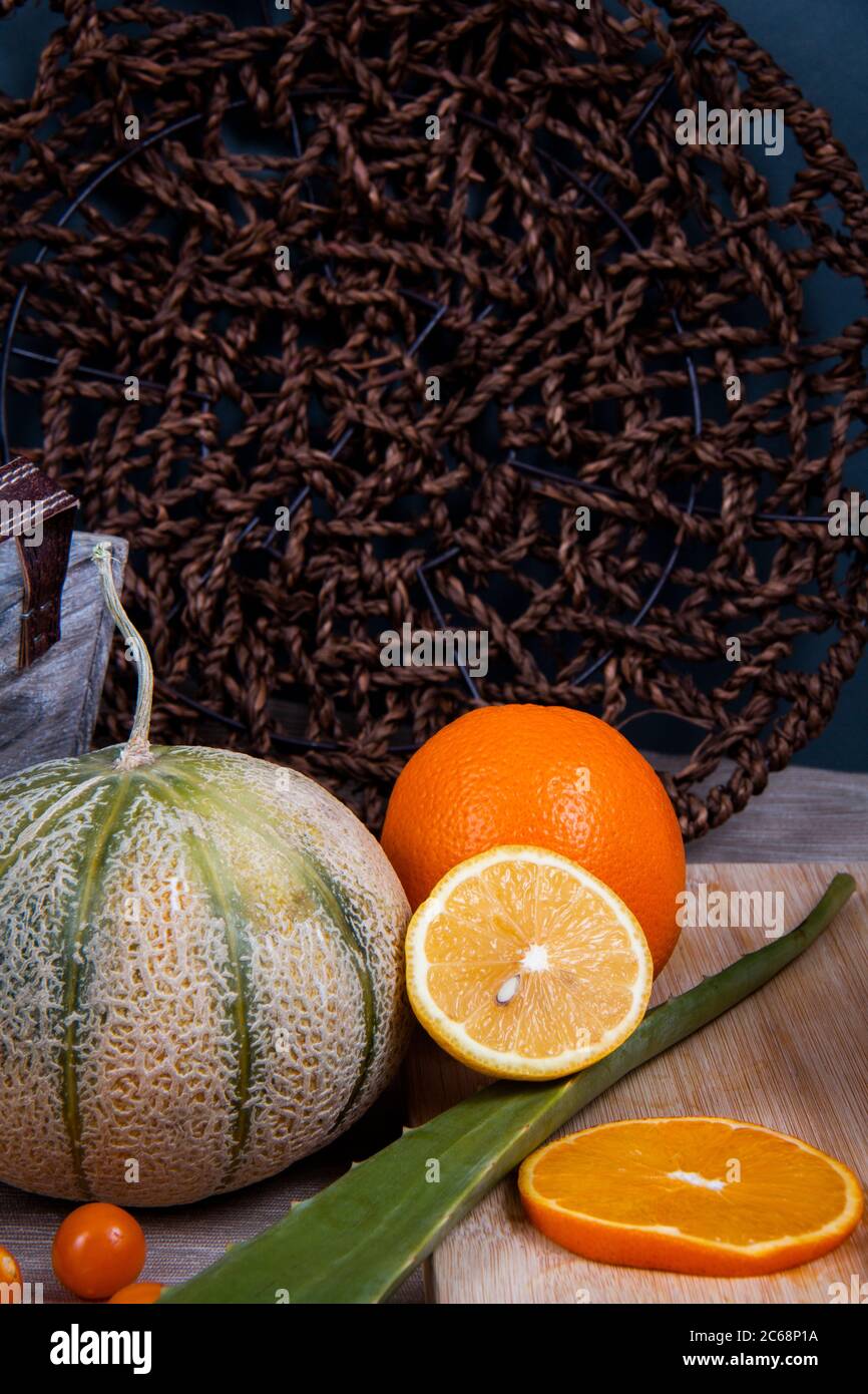 Obst-Fotografie von einigen essbaren Früchten: Orange, Honigmelone, chinesische Laternen Früchte und ein grünes Blatt Aloe Vera, vor einem gewebten flachen Korb Stockfoto