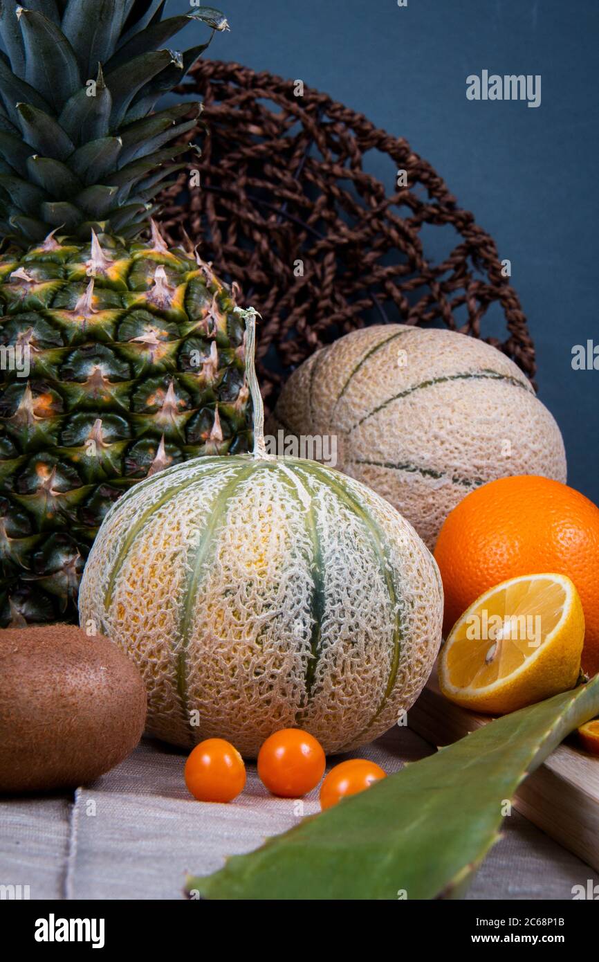 Obst-Fotografie von einigen essbaren Früchten: Orange, Honigmelone, chinesische Laternen Früchte und ein grünes Blatt Aloe Vera, vor einem gewebten flachen Korb Stockfoto
