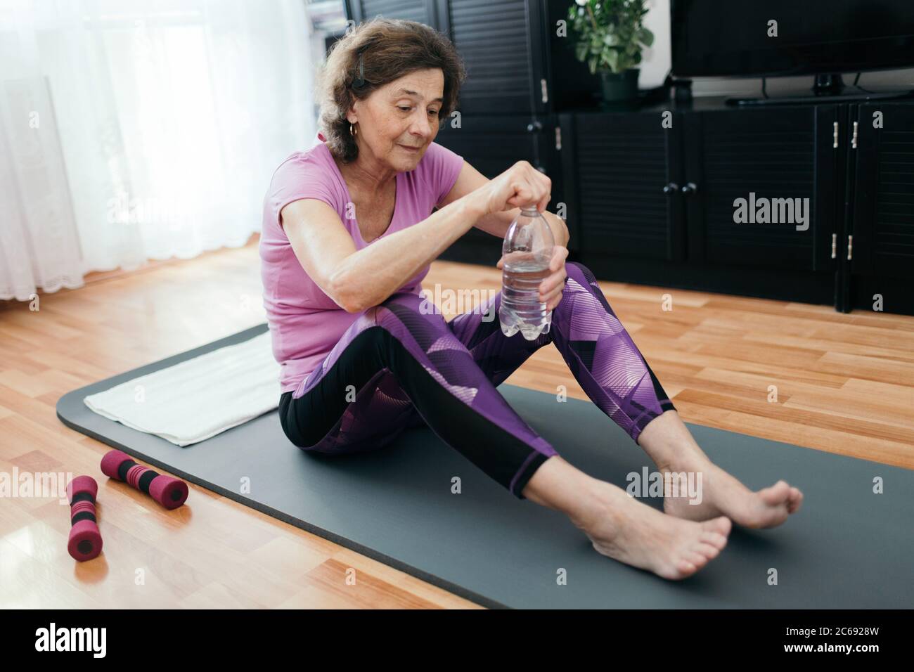 Ältere Frau wird nach dem Training zu Hause Wasser trinken. Müde ältere Frau in lila Sportkleidung sitzt auf Fitness-Matte Öffnung Flasche mit Wasser Stockfoto