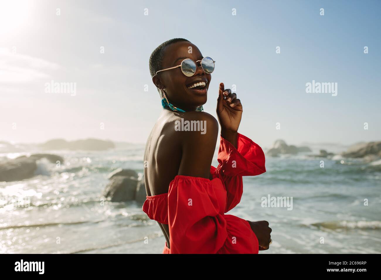 Attraktive Frau in roten Kleid tanzen am Strand. Afrikanische Frau trägt rote Sonnentress und Sonnenbrille Spaß am Strand. Stockfoto