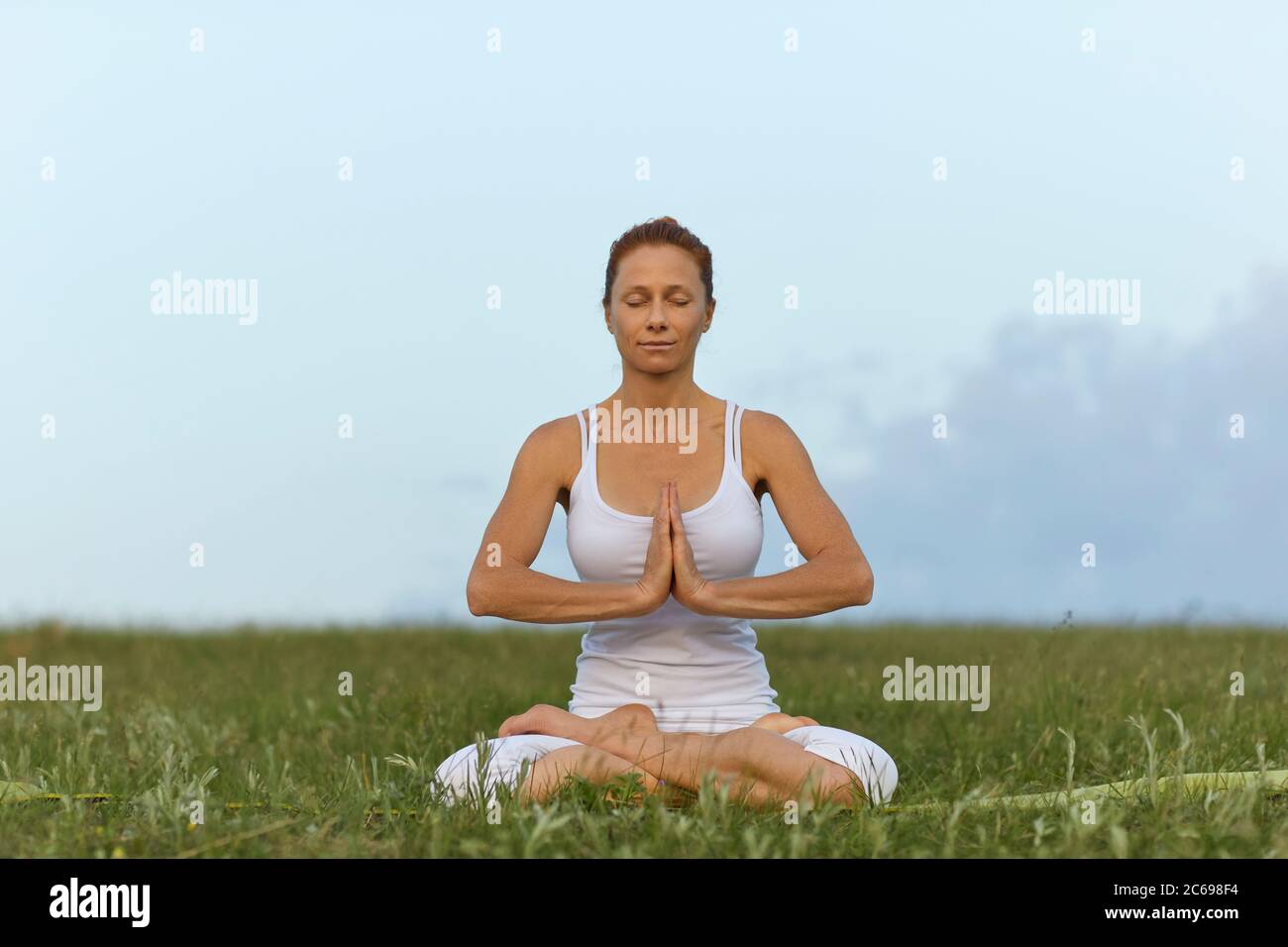 Frau Yoga in der Natur. Ein Mädchen in weißen Kleidern praktiziert Meditation, während es in einer Lotusposition auf dem Gras gegen den Himmel sitzt. Stockfoto
