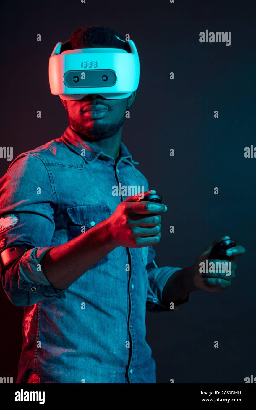 Genießen neue Erfahrung. Hübscher junger afrikanischer Mann im VR-Headset Gestik und lächelnd, während gegen Rot und Blau Dual Color Licht auf Dunkle b Stockfoto