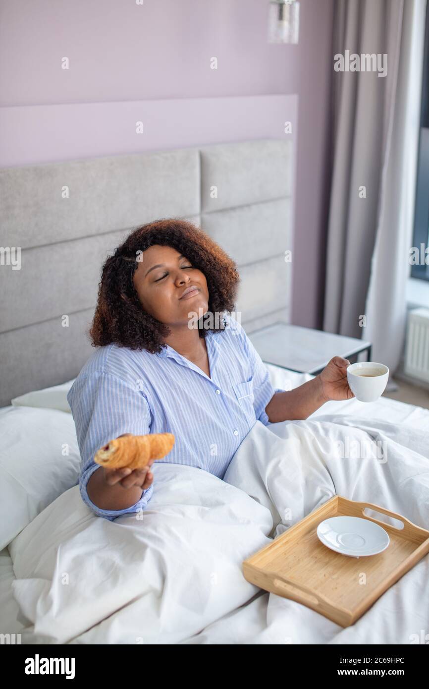 Lustige Frau mit geschlossenen Augen hält eine Tasse Tee und Croissant im Schlafzimmer. Close up Seite Ansicht Foto Stockfoto