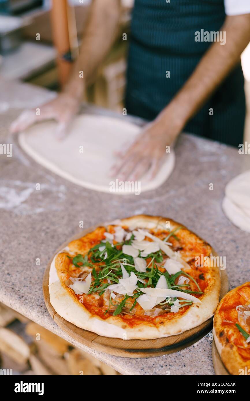 Fertige Pizza mit Käse und Rucola vor dem Hintergrund des Küchenchefs Pizza, rollt den Teig auf dem Tisch. Stockfoto