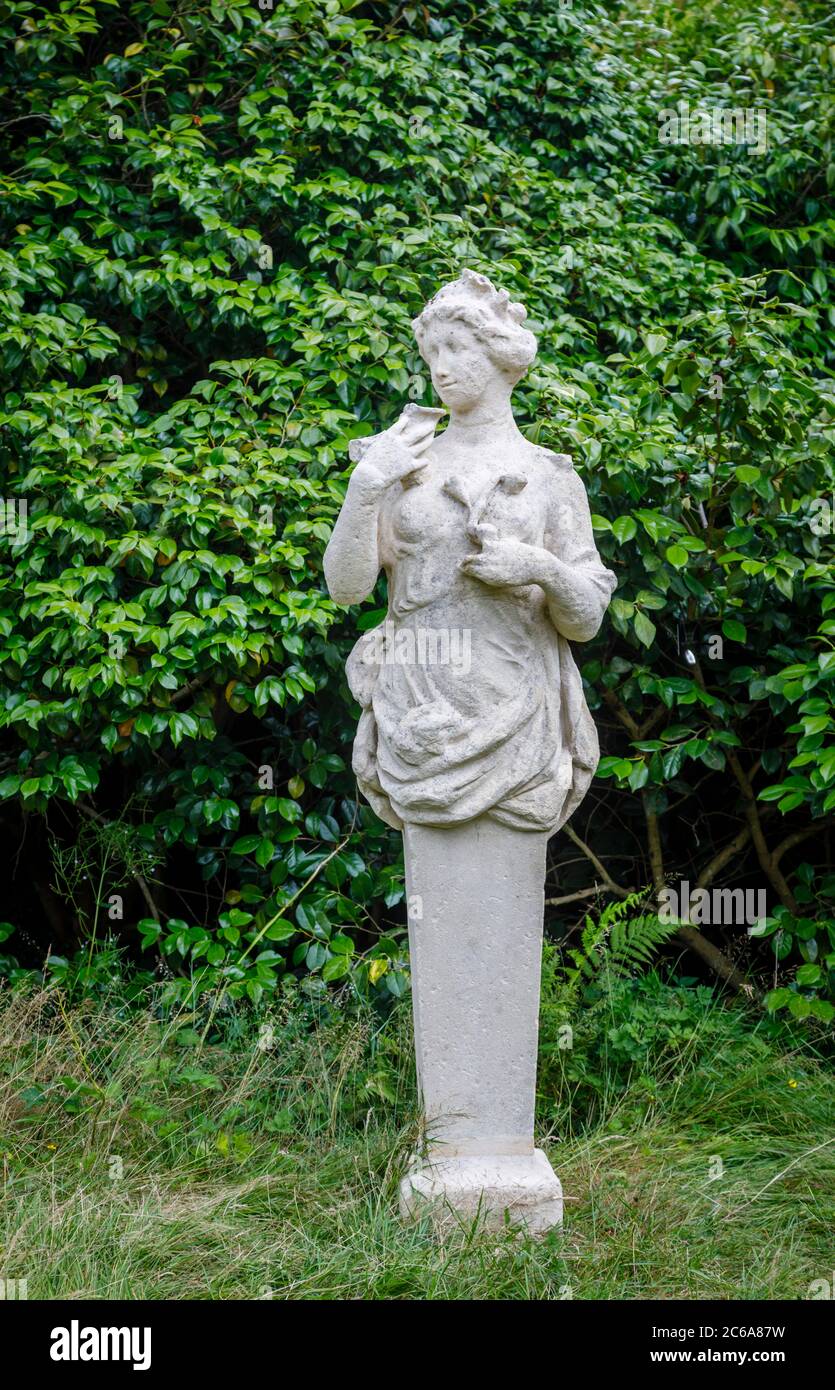 Klassische leicht erodierte Vintage-Steinstatue einer weiblichen Nymphe oder Dryade, die in einem Garten im Südosten Englands vor einem grünen Heckenhintergrund steht Stockfoto