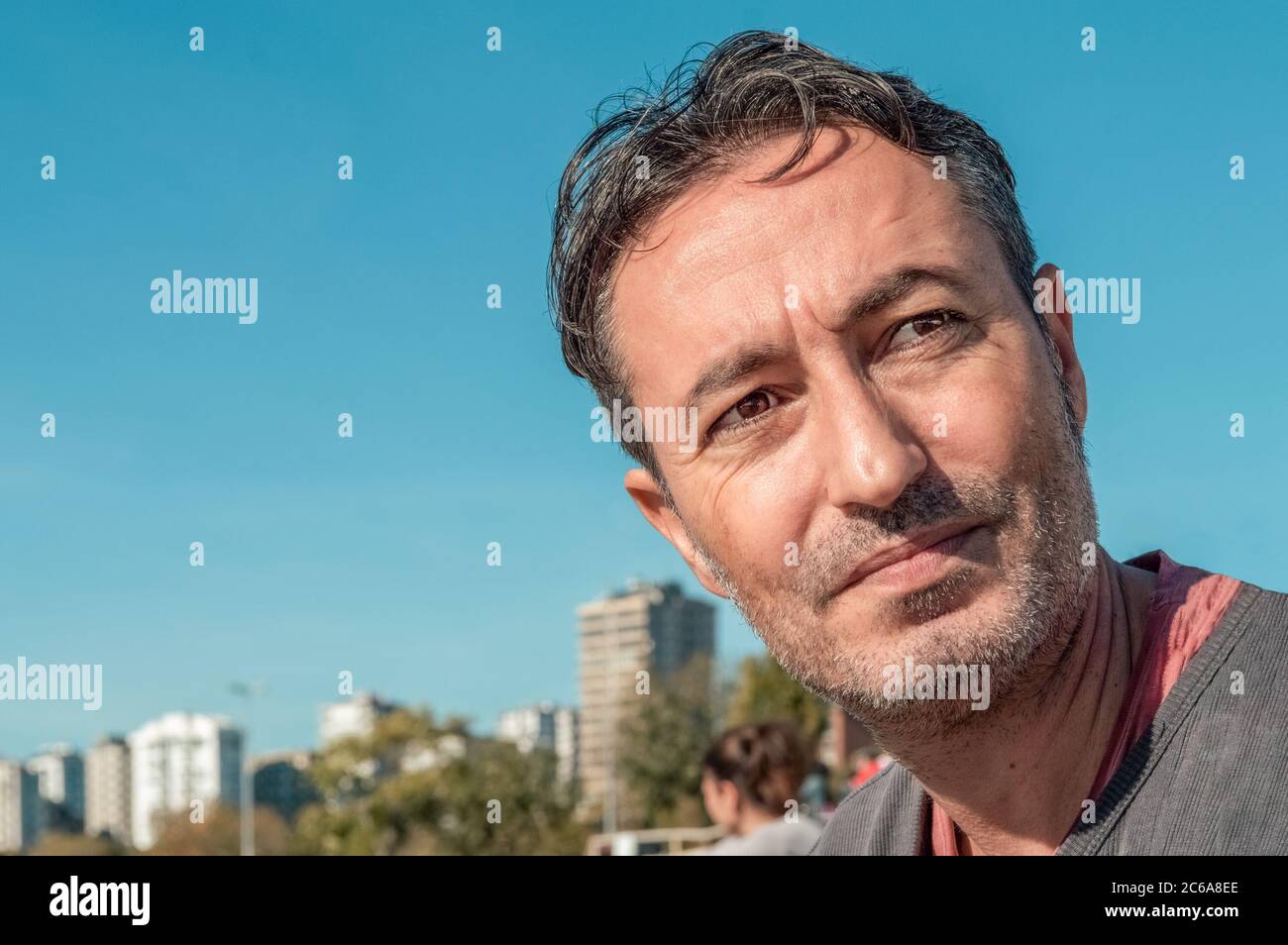 Outdoor-Porträt eines schönen Brunet Mann im Park mit blauem Himmel und städtischen Hintergrund. Verbringen Sie Zeit am Wochenende. Emotionaler Ausdruck. Stockfoto