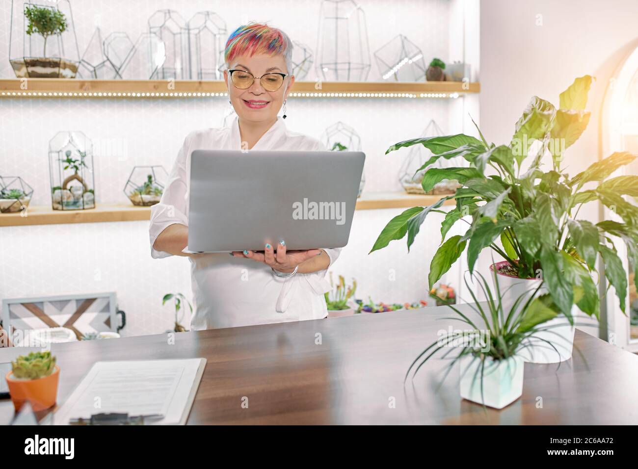 Schöne kaukasische ältere Frau in weißen Bademantel scharf auf Flora gekleidet. Stellen Sie sich in einem hellen Raum voller Pflanzen auf und betrachten Sie den Bildschirm des Laptops Stockfoto