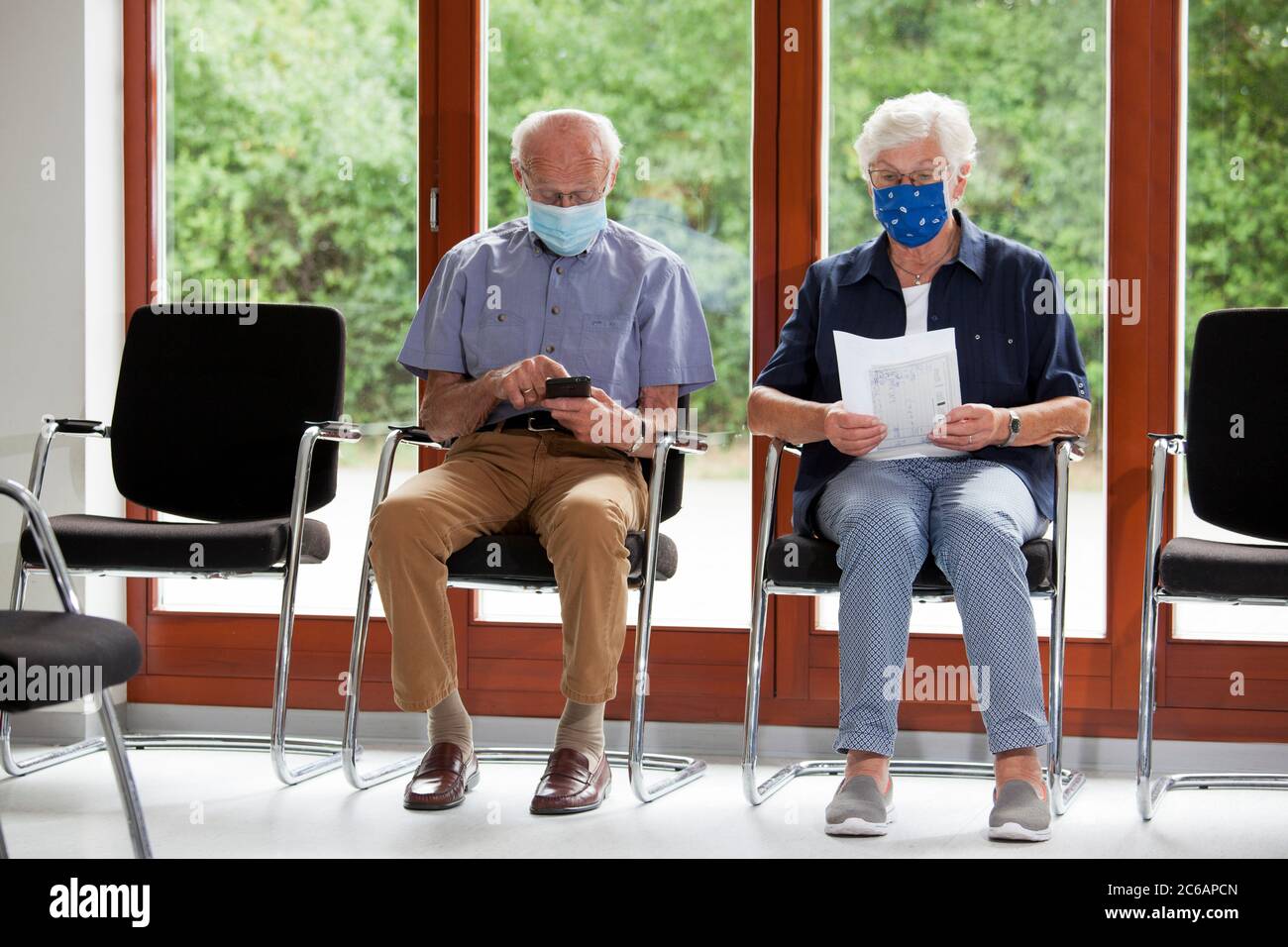 Seniorenpaar sitzt mit Gesichtsmasken in einem hellen Wartezimmer eines Krankenhauses oder eines Büros - der Mann schaut auf ein Smartphone Stockfoto