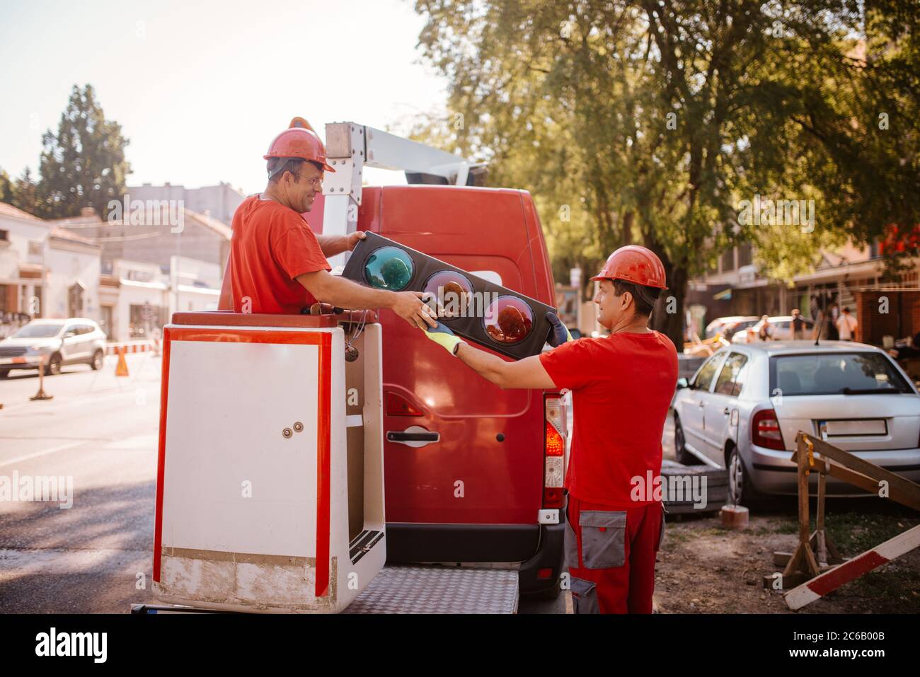 Ein Kaukasischer Arbeiter mittleren Alters in roter Uniform und Helm passiert eine Ampel zu einem anderen Arbeiter. Einrichten von Ampeln auf der Straße in prograe Stockfoto