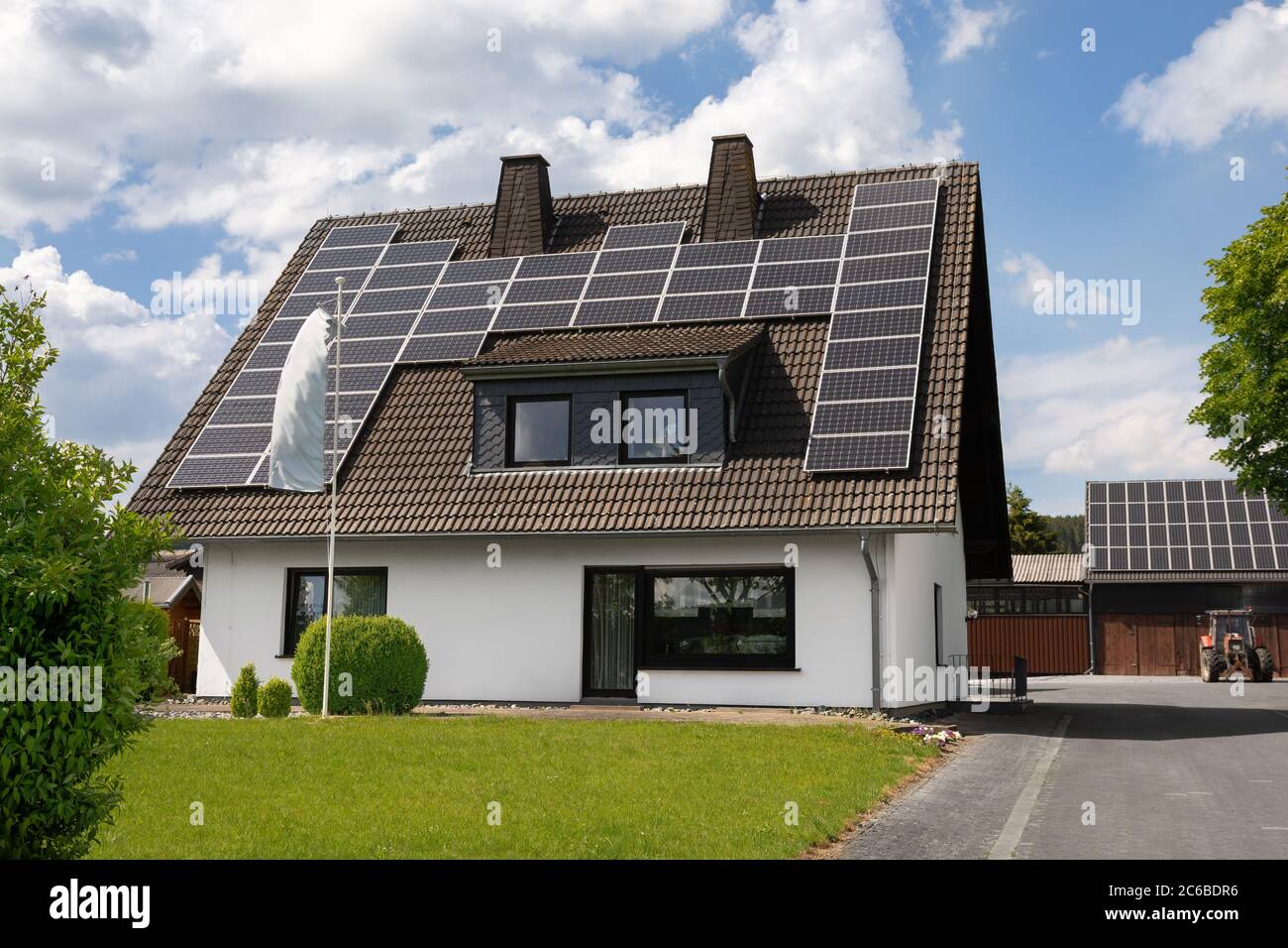 Ein Haus mit Solarzellen auf dem Dach. Ein Bauernhof wird mit Solarenergie betrieben. Im Hintergrund sind Stallungen. Haus in grüner Natur. Blauer Himmel mit Wolken Stockfoto
