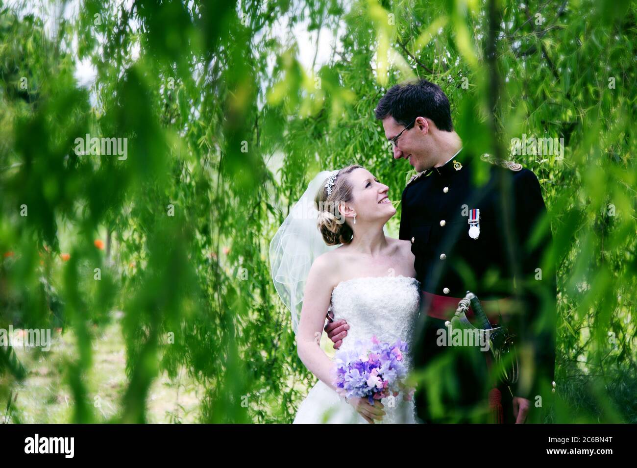 Braut und Bräutigam, in Militäruniform mit Schwert, lächelnd unter einer Weide an ihrem Hochzeitstag Stockfoto