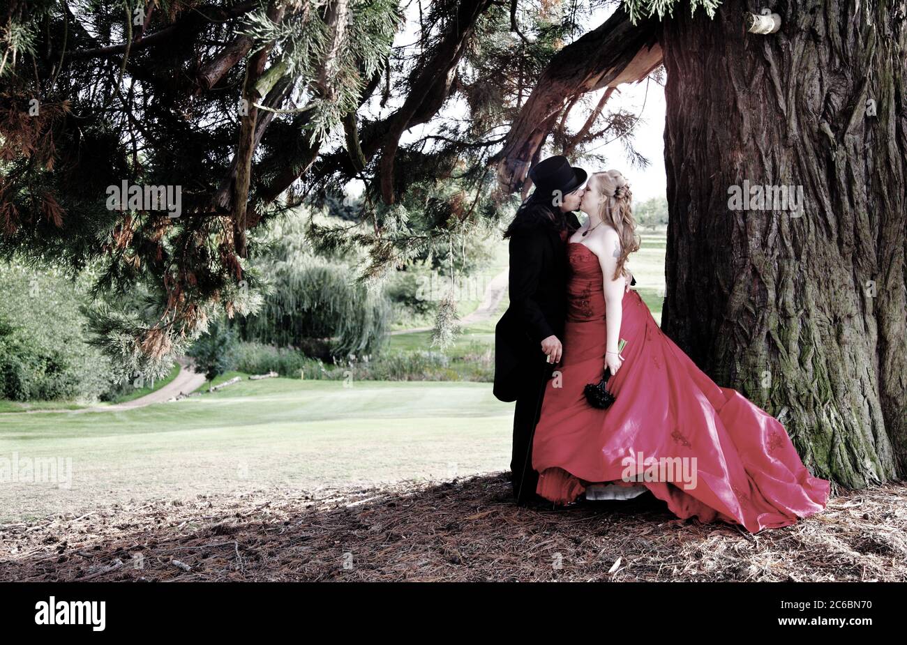Bräutigam Dreißiger & Braut Zwanziger in Gothic Hochzeitsmode mit rotem Kleid, Gothic Zylinderhut, Schwänze & Stock. Paar küssen unter einem Baum. Stockfoto