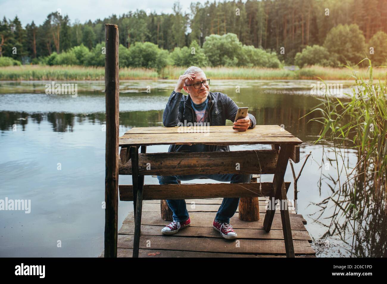 Minsk, Weißrussland - 13. Juni 2020: Mann auf einem hölzernen Pier am Fluss in der Natur - nebliger Morgen - mobile Kommunikation für Fernarbeit Stockfoto