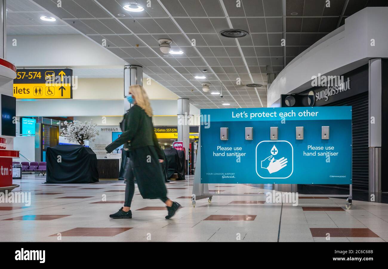 Eine Frau mit Gesichtsmaske geht während der weltweiten Coronavirus-Pandemie 2020, England, an einer Station für Händedesinfektionsmittel am Flughafen Gatwick vorbei Stockfoto