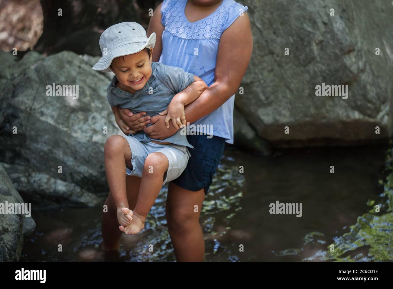 Große Schwester hält ihren kleinen Bruder über dem kalten Wasser aus einem Teich, während er nervös ist. Stockfoto