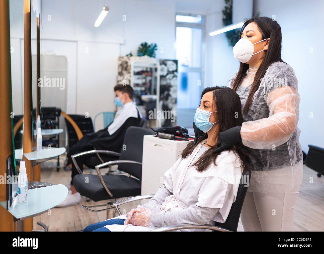 Friseur mit einer Schutzmaske und Handschuhen, die das Haar eines maskierten Kunden messen, der auf einem Stuhl vor einem Spiegel in einem Friseursalon sitzt Stockfoto