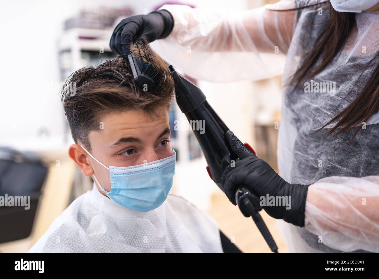 Friseur Trocknen von Haar mit einem elektrischen Trockner zu einem Jungen mit einer Schutzmaske sitzt auf einem Stuhl in einem Friseursalon Stockfoto