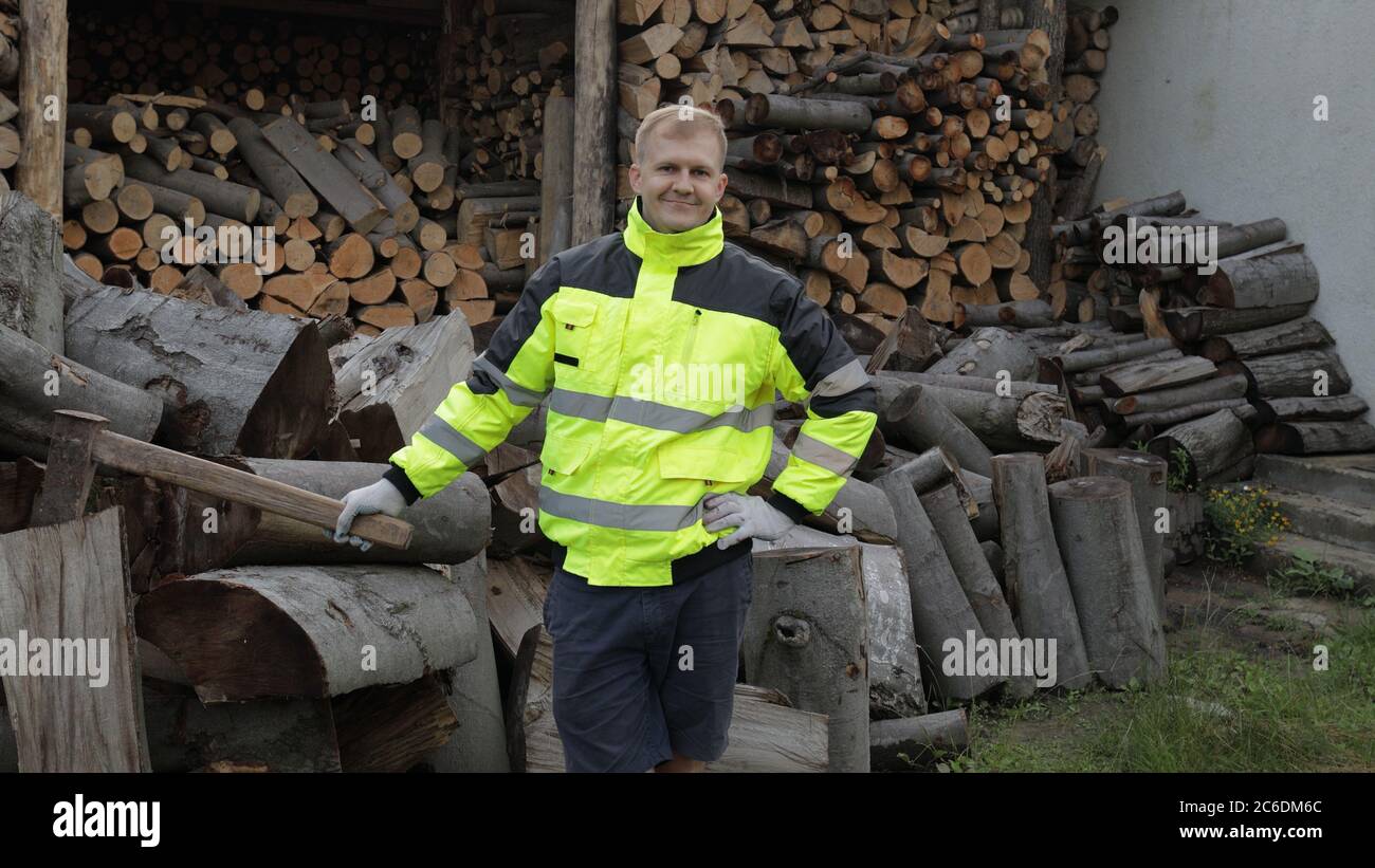 Holzfäller posiert in grüner Jacke mit reflektierenden Streifen und hält große Axt. Bereit für die Arbeit. Holzfäller stehend und Kamera anblickend. Gesägte Holzstämme, Brennholz Hintergrund. Sägewerk, Holzernte Stockfoto