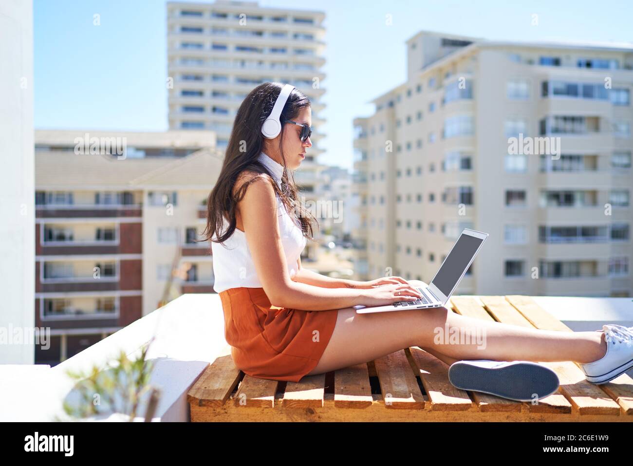 Junge Frau mit Kopfhörern mit Laptop auf sonnigen städtischen Balkon Stockfoto