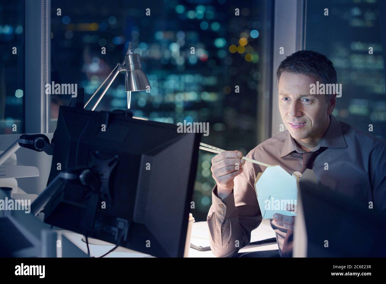 Geschäftsmann, der abends im Büro Essen zum Mitnehmen am Laptop isst Stockfoto