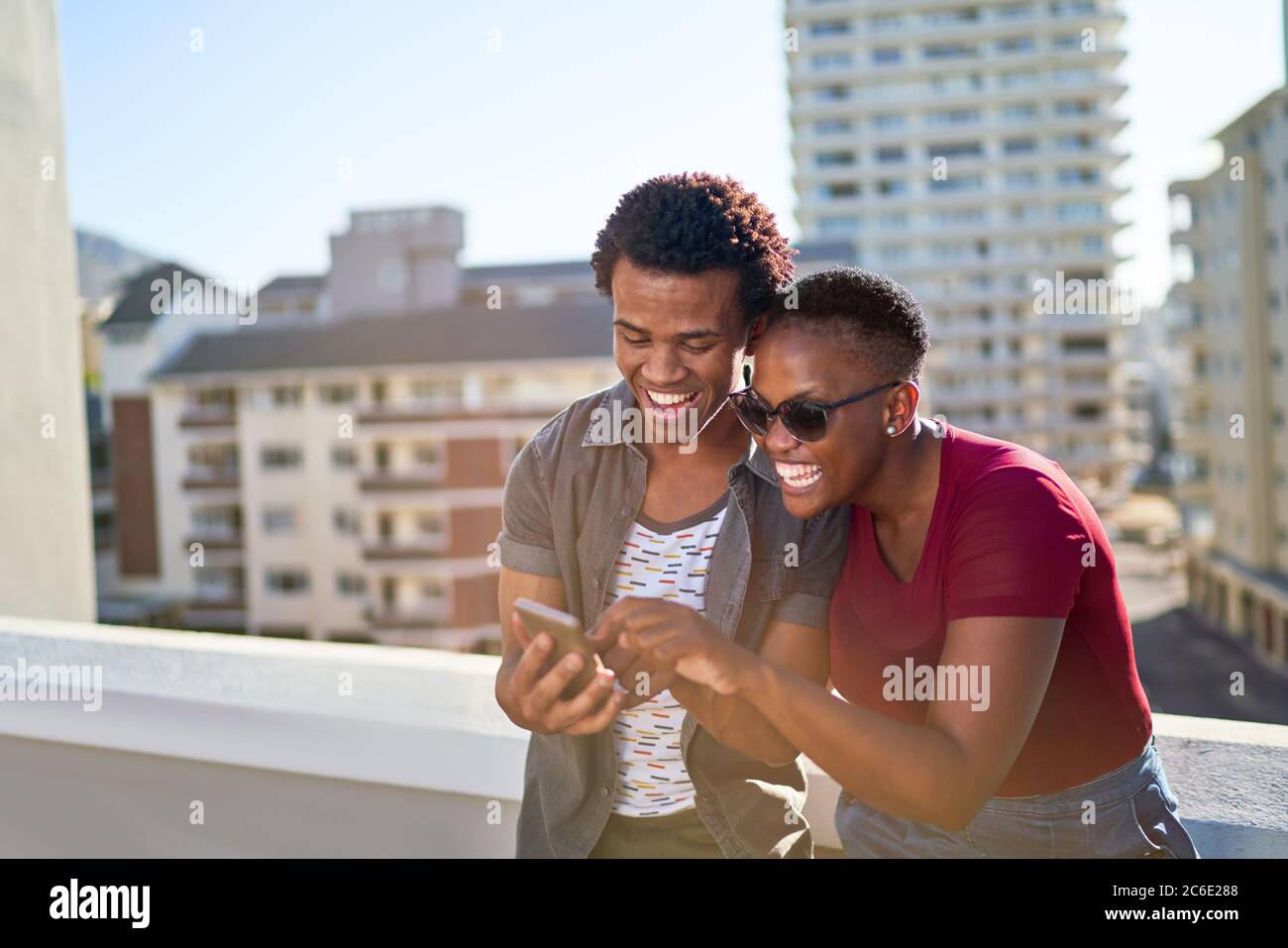 Glückliches junges Paar mit Smartphone auf sonnigen städtischen Dachbalkon Stockfoto